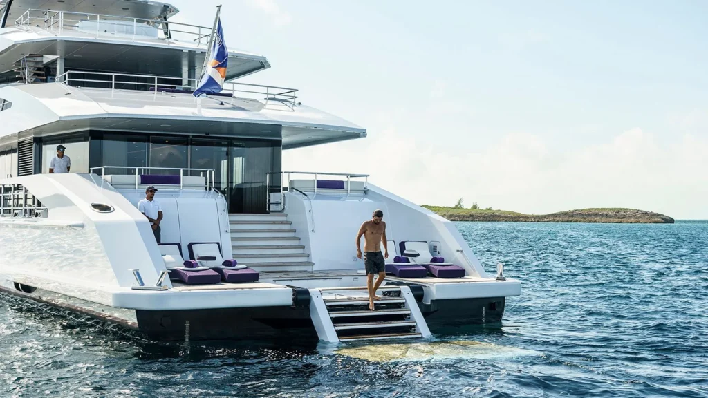 Vista de popa de un superyate Heesen de color blanco y 50 metros de eslora anclado en las aguas de Porto Cervo, Costa Esmeralda, Cerdeña. La imagen muestra la zona del club de playa con los miembros de la tripulación vestidos con uniformes blancos de pie en la cubierta y un invitado bajando las escaleras hacia el mar. La zona de descanso está amueblada con tumbonas con característicos cojines morados que contrastan con el color del barco.