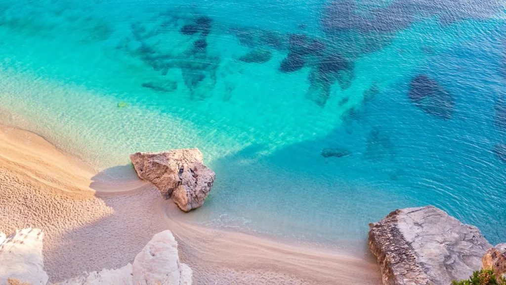 A stunning aerial view of Cala Goloritzè in the Gulf of Orosei, Sardinia. The image shows the iconic white pebble beach and the towering limestone cliffs meeting the transparent turquoise and deep blue waters of the Mediterranean, highlighting the unique geological formations of this UNESCO site.