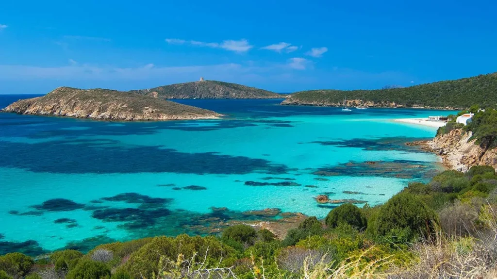 A wide coastal view near Sant’Antioco in southwestern Sardinia, showing vibrant turquoise waters and rocky islands in the distance. The image features a small white sandy beach on the right and green Mediterranean scrub covering the hillsides in the foreground.