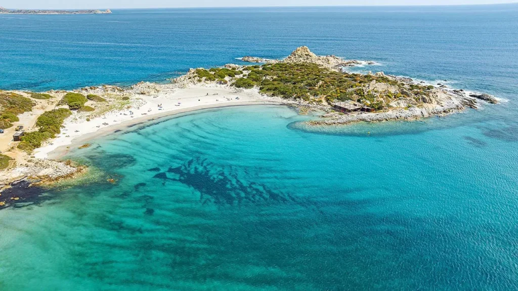 A stunning aerial view of Punta Molentis beach near Villasimius on the southeastern coast of Sardinia. The image shows the characteristic crescent of white sand and shallow turquoise waters of the Capo Carbonara Marine Protected Area, surrounded by rocky cliffs and Mediterranean scrub.