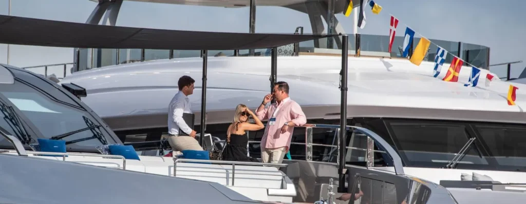 A professional yacht broker in a white shirt and tan trousers discusses the buying process with a couple on the deck of a luxury vessel in Porto Cervo, Costa Smeralda, Sardinia. The group is engaged in an expert consultation surrounded by high-end yachts and nautical flags.