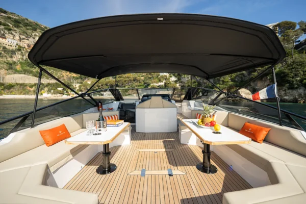 Outdoor aft dinette of the BB Yachts Panarea 44 featuring two black tables, beige sofas, and orange pillows under a black bimini top in Sardinia.