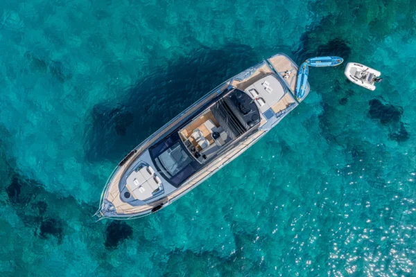 An aerial top-down view of a grey Cranchi 60 ST yacht anchored in turquoise, transparent water with a visible rocky sea floor, typical of the coastal areas around Porto Cervo, Costa Smeralda, Sardinia.