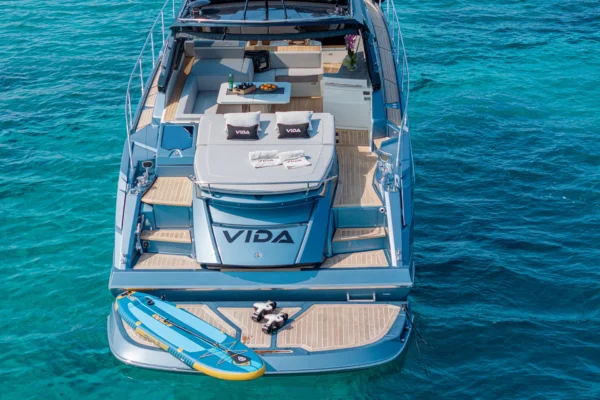A direct view of the Cranchi 60 ST stern from behind, showing the wide teak swim platform with an inflatable paddleboard resting on it, leading up to the cockpit area under the bright sky of Porto Cervo, Costa Smeralda, Sardinia.