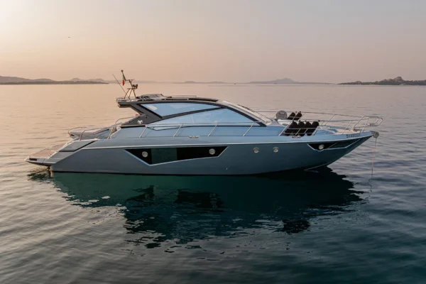 A rear side profile view of a Cranchi 60 ST at anchor during sunset, with its hull reflected in the calm water and a soft orange glow on the horizon behind the silhouettes of Porto Cervo, Costa Smeralda, Sardinia.