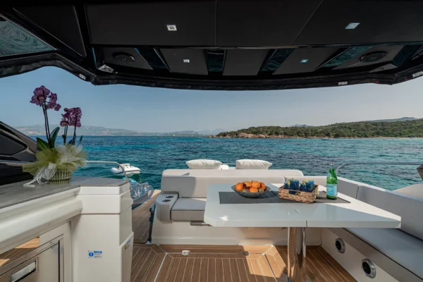 An elevated view looking forward onto the rear cockpit lounge of the Cranchi 60 ST. A wooden table is surrounded by light grey u-shaped seating. On the table sits a fruit bowl and a wine bottle. To the left, a purple orchid in a decorative planter is visible. The view extends past the seating to clear blue sea and a rugged, green coastline.