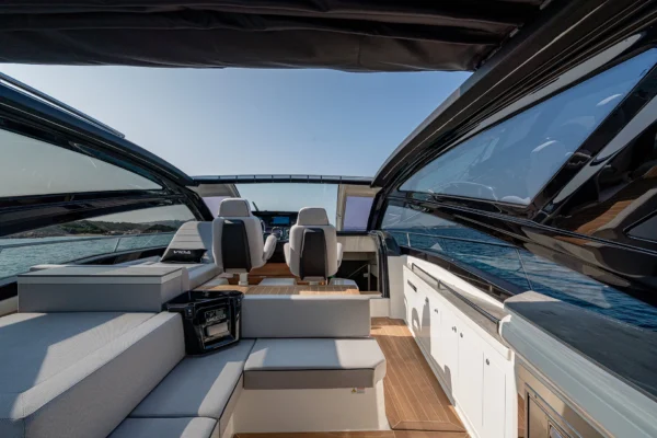 A wide, rear-facing perspective from within the cockpit area of the Cranchi 60 ST, looking towards the helm. The modern steering station with dual seats is on the left, while on the right is a dining area with a table and bench seating, all covered by a sleek, openable sunroof structure.