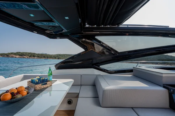 A close-up view of the rear cockpit seating area on a Cranchi 60 ST yacht. Light grey cushions line a banquette seat. In front of it is a table set with a woven fruit basket and two ceramic items. The background shows the clear blue water of the open sea under a sunny sky.