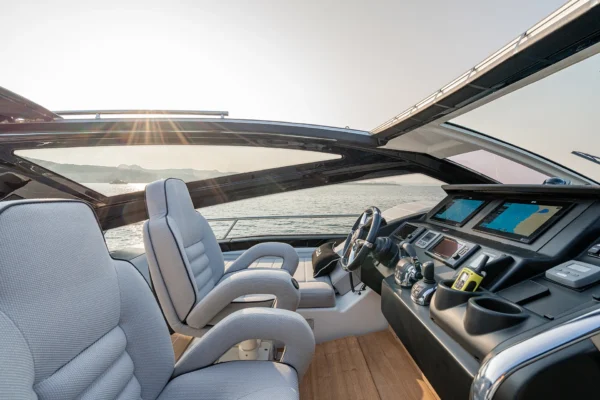 A detailed interior view of the modern helm station on a Cranchi 60 ST yacht. Two grey leather pilot seats are positioned behind a dark dashboard with digital display screens, a steering wheel, and throttle controls. Teak flooring covers the deck. The forward windscreen looks out onto water and distant land under a bright sky.
