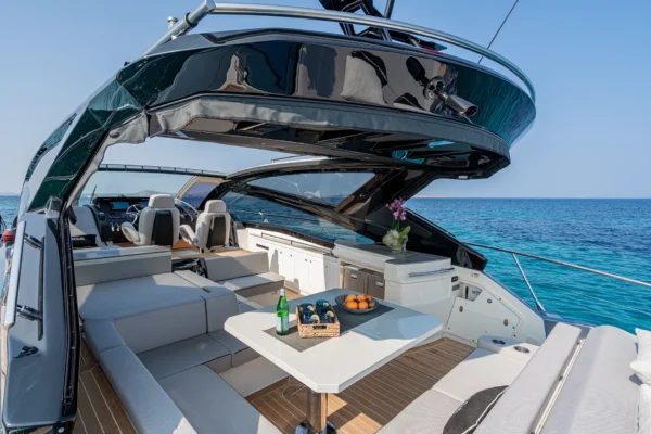 A rear-view looking forward over the open cockpit of the Cranchi 60 ST. A large, dark composite arch-structure supports an integrated sunroof mechanism. Below it is a spacious dining and seating area with a table. Teak decking is visible throughout, leading towards the helm station in the distance, set against a backdrop of blue ocean and sky.