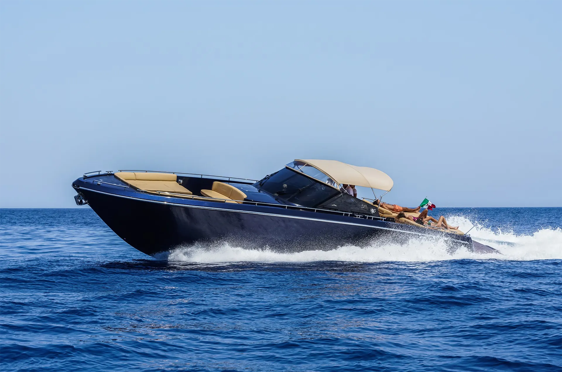 continental-50-open-yacht-profile-navigation A side-profile photograph of the dark blue Continental 50 open yacht cruising at high speed on blue water, showing its low profile, beige cockpit cushions under a matching canvas top, and a prominent white wake at the stern.