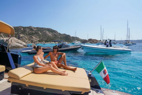 A close-up shot from the stern sunbed of the yacht showing a man and a woman sitting and talking, with an Italian maritime flag in the foreground and other yachts anchored near a rocky coastline in the background.