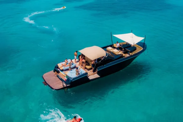 High-angle view of the Continental 50 yacht deck crowded with guests at anchor. The setup shows a large wooden swim platform at the stern, extended bimini awnings shading the seating areas, and a white sunshade rigged over the bow.