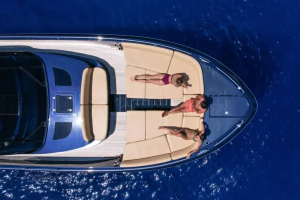 Inquadratura zenitale della prua dello yacht Continental 50. Tre persone sono stese comodamente sugli ampi cuscini prendisole beige, prendendo il sole. La coperta in teak contrasta con la tuga blu scuro e il blu intenso del mare circostante.