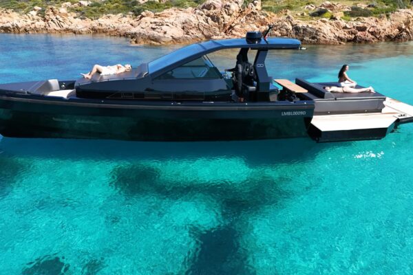 Profile view of the black Seainfinity T5 walkaround yacht floating on crystal-clear sea water next to the rocky coast of Sardinia, highlighting its aggressive lines, tinted glass hardtop, and extended side terraces.