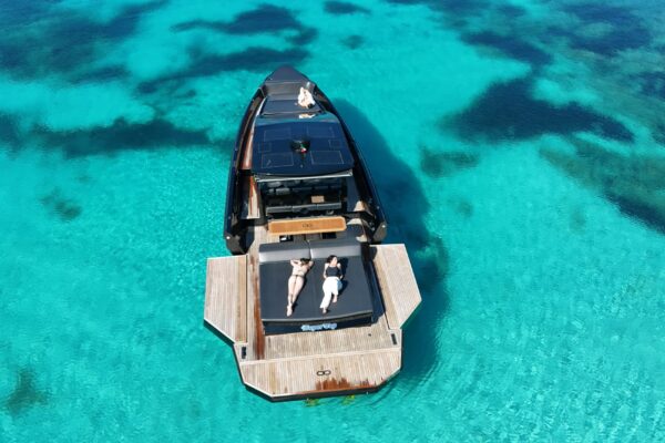 A top-down aerial drone shot of the black Seainfinity T5 motor yacht, showcasing the massive cockpit space created by both port and starboard hydraulic side terraces fully extended on the clear sea of Costa Smeralda, Sardinia.