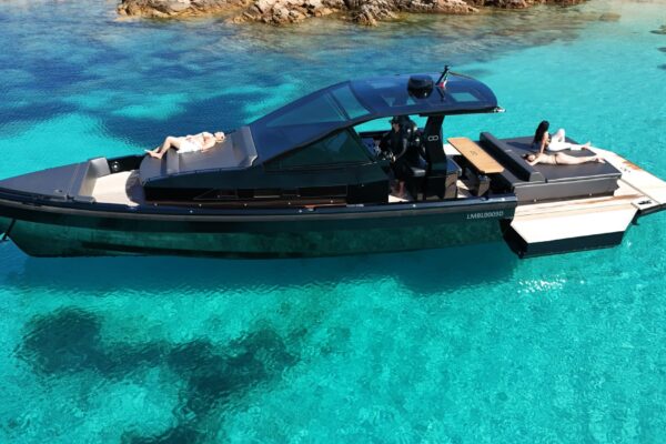 A broadside drone photograph of the black Seainfinity T5 luxury yacht at anchor in the turquoise water of Porto Cervo. A guest relaxes on the forward sunbed while others enjoy the aft deck lounge with the starboard hydraulic terrace fully opened.