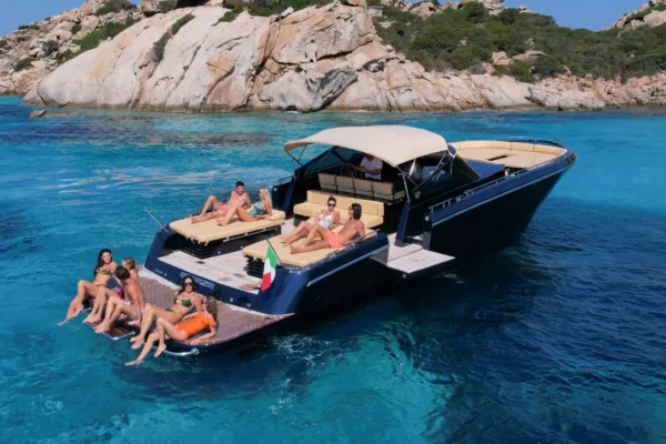 Un gruppo di amici si diverte e si rilassa a poppa di uno yacht Continental 50 ormeggiato vicino a una scogliera rocciosa. Alcune persone sono sedute con i piedi in acqua, altre sono stese sui prendisole, con lo skipper visibile al posto di comando sotto il tendalino.