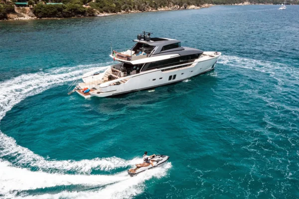 Aerial view of the Sanlorenzo SX88 anchored in clear water with a guest riding a Sea-Doo personal watercraft in a circular path around the yacht, leaving a white foam wake.