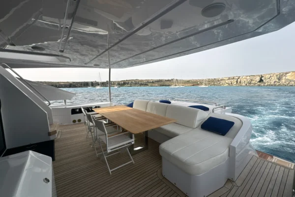 Covered main deck aft cockpit of the Amer F100 superyacht featuring a wooden dining table, white lounge seating, and teak flooring overlooking the sea.