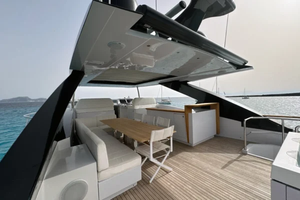 Shaded flybridge deck of the Amer F100 superyacht featuring a wooden dining table, white seating, teak flooring, and a wet bar area under the hardtop.