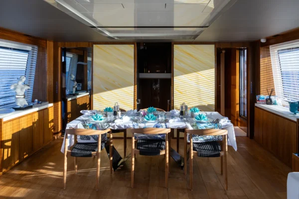 Elegant formal dining area inside the Amer F100 superyacht, fully set with marine-themed decor and blue napkins, surrounded by warm wood paneling, decorative backlights, and large side windows.