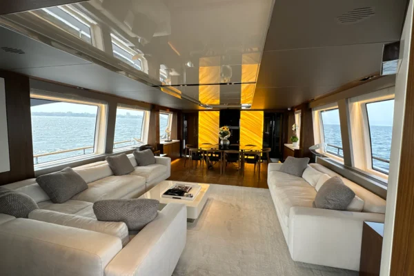 Expansive main deck saloon of the Amer F100 superyacht featuring facing white sofas, panoramic ocean view windows, and a forward formal dining area highlighted by a striking backlit onyx wall.
