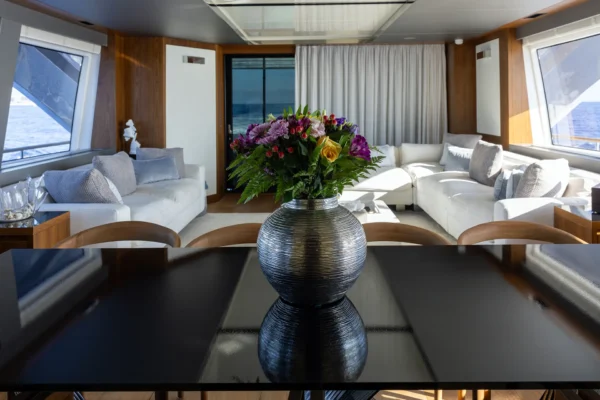 Close-up of a silver floral vase on a dark dining table, revealing the modern main saloon of the Amer F100 superyacht with white sofas, warm wood paneling, and panoramic ocean view windows.