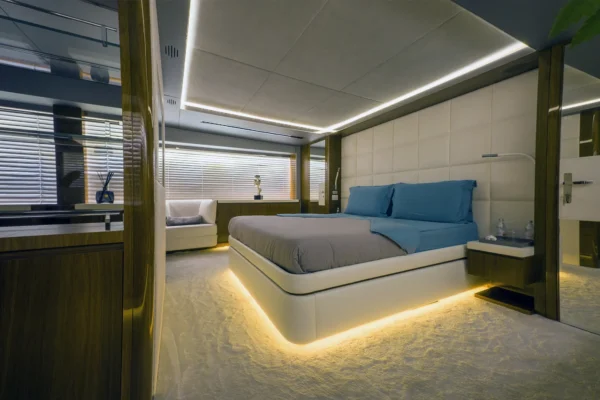 Spacious master stateroom on the Amer F100 superyacht highlighting a large central bed with an illuminated base, a white padded headboard, polished wood paneling, mirrored wardrobes, and elegant modern ceiling lighting.