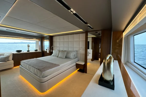 Luxurious master stateroom on the Amer F100 superyacht showcasing a large central bed with LED under-lighting, a white quilted headboard, elegant wood paneling, and wide side windows offering panoramic sea views.