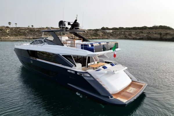 Elevated aft quarter view of the Amer F100 superyacht anchored in calm waters, highlighting the multi-level deck spaces, swim platform, and dark blue hull.