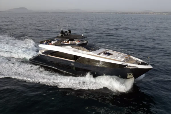 Aerial three-quarter bow view of the Amer F100 superyacht cruising dynamically through the open sea, displaying its distinctive dark blue hull, white wake, and spacious foredeck equipped with large sunpads.