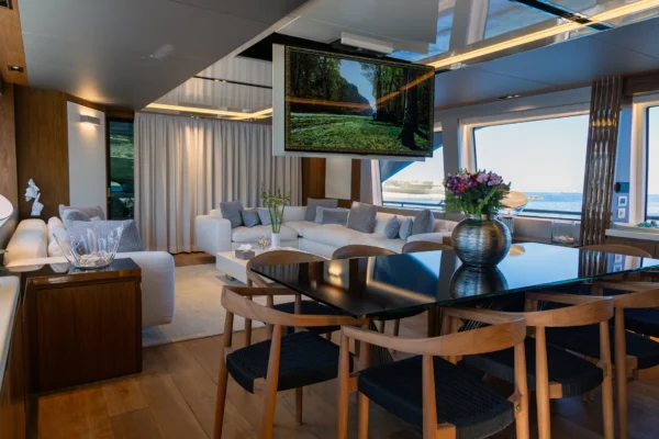 Spacious main deck saloon of the Amer F100 superyacht featuring warm wooden flooring, a formal dining space, plush white lounge seating, and a large modern drop-down television screen.