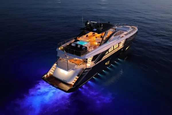 Dramatic aerial stern view of the Amer F100 superyacht cruising at twilight, featuring vibrant blue underwater LED lights illuminating the wake and warm exterior deck lighting highlighting the flybridge jacuzzi and aft cockpit.