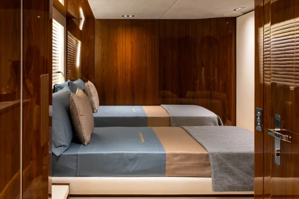 View looking into a twin guest cabin on the Amer F100 superyacht through a polished wood doorway, displaying two single beds dressed in soft blue and grey linens against rich wooden walls.