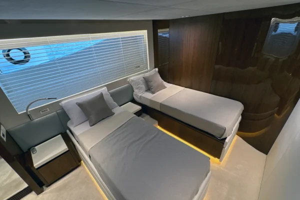 Modern twin guest cabin on the Amer F100 superyacht featuring two single beds with illuminated bases, warm high-gloss wood paneling, individual reading lights, and a window with white privacy blinds.