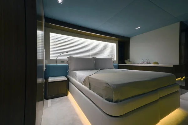 Elegant VIP double guest cabin on the Amer F100 superyacht featuring a plush bed with warm under-bed LED lighting, soft blue and grey accents, dark wood cabinetry, and a large window with privacy blinds.