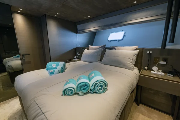 Interior view of a luxury double guest cabin on the Sanlorenzo SX88 charter yacht, featuring a neatly made bed accented with rolled teal towels, modern grey wood finishes, and a classic porthole window.