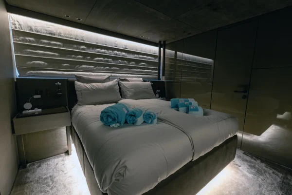 Sophisticated double cabin interior on the Sanlorenzo SX88, showcasing a spacious bed decorated with bright teal towels, atmospheric under-bed LED lighting, and sleek, dark glossy wall paneling.