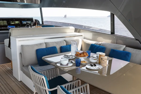 Covered flybridge deck on the Sanlorenzo SX88 featuring a modern, glossy dining table set for breakfast with woven chairs and blue accent pillows, located directly behind the yacht's high-tech helm station with digital navigation screens.