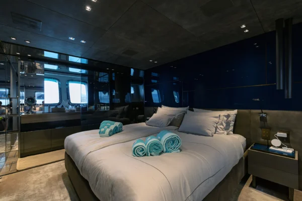 Luxurious master suite interior aboard the Sanlorenzo SX88 charter yacht, featuring a spacious double bed decorated with rolled teal towels, high-gloss dark blue wall paneling, and a modern glass partition revealing the en-suite bathroom.