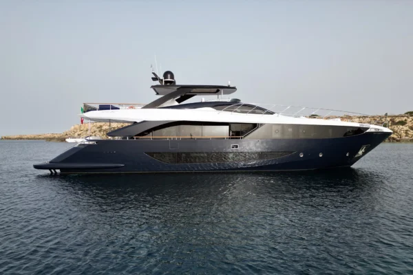 A side profile view of the blue and white Amer F100 superyacht anchored in a calm bay near a rocky coast.