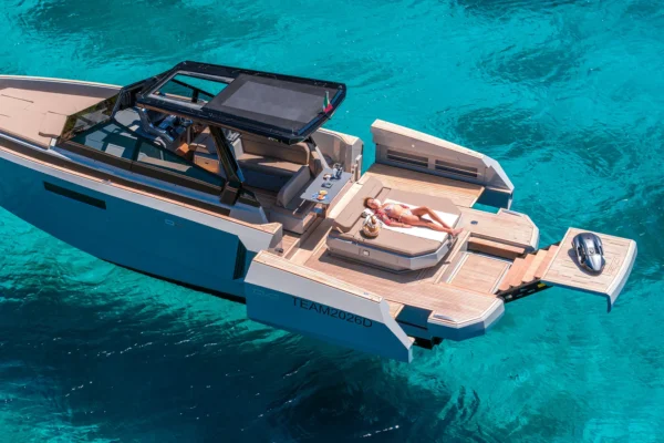 Elevated aerial view of the EVO 43 WA motor yacht anchored in bright turquoise water. The image clearly displays the fully expanded side platforms and the extended hydraulic stern platform holding a water toy. A guest is sunbathing on the large central aft sunpad, highlighting the expansive open deck, the walk around layout, and the open sunroof on the black hardtop.