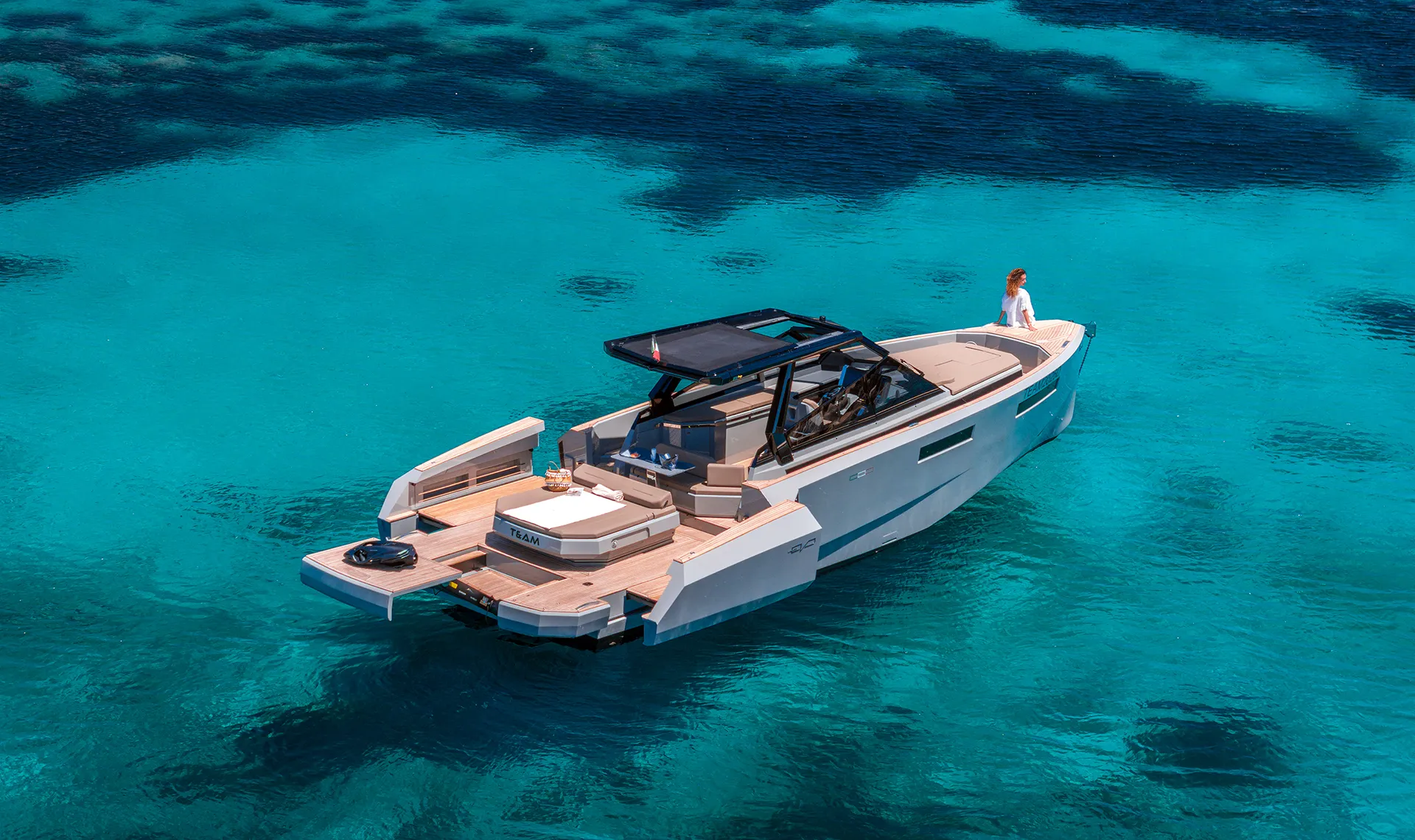 evo-43-wa-yacht-charter-expandable-platforms-aerial Aerial view of the EVO 43 WA luxury motor yacht anchored in clear turquoise waters. The vessel is shown with its expandable side platforms fully open, maximizing the aft deck space, with the walk around configuration clearly visible and a guest relaxing on the bow.